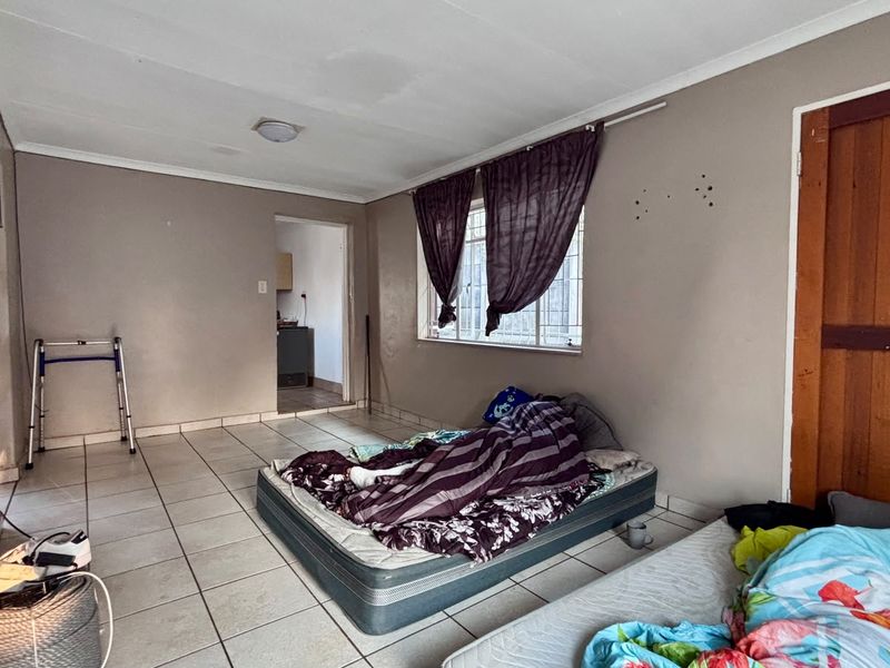 A bedroom featuring tiled flooring, a window with security bars, and a wooden door, requiring cosmetic updates to the walls and ceiling.