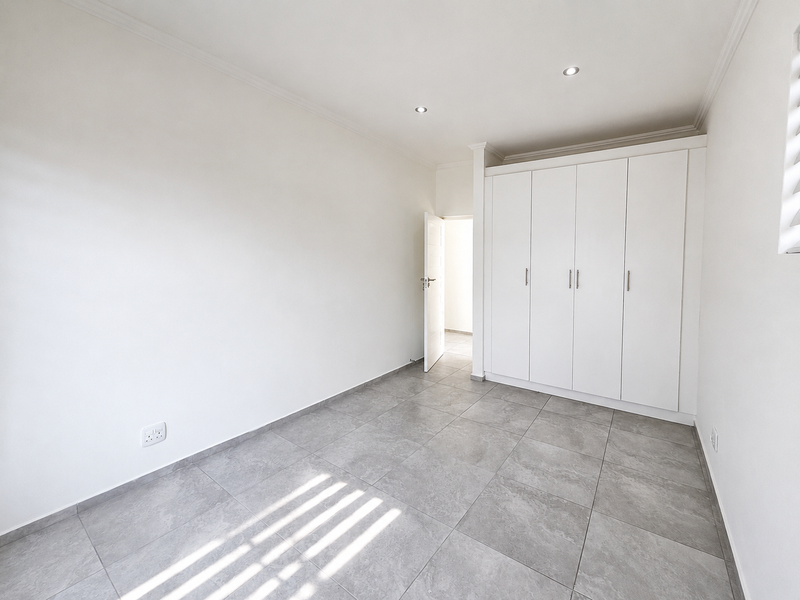 A bright, empty bedroom featuring modern grey floor tiles, built-in white cupboards, and recessed ceiling lighting.