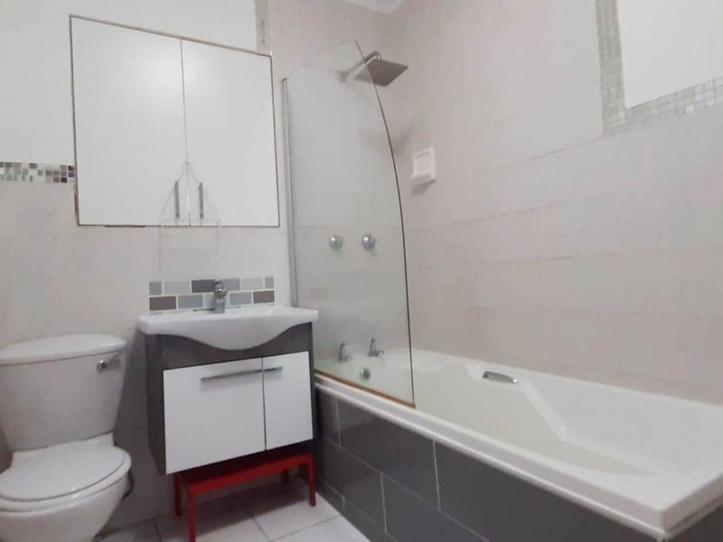 A clean, functional bathroom featuring a built-in vanity, a bathtub with a glass shower screen, and neutral wall tiling.