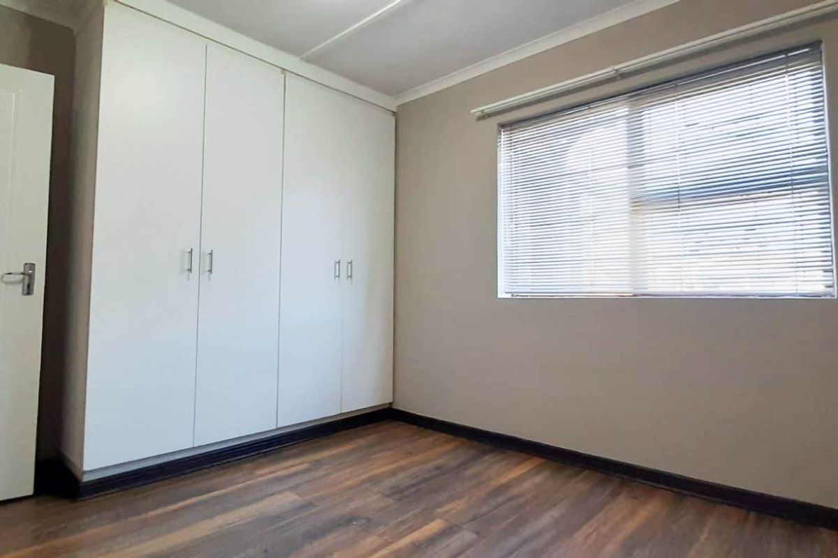 A clean, empty bedroom featuring modern laminate flooring, built-in white cupboards, and venetian blinds, offering a neutral space ready for personalization.