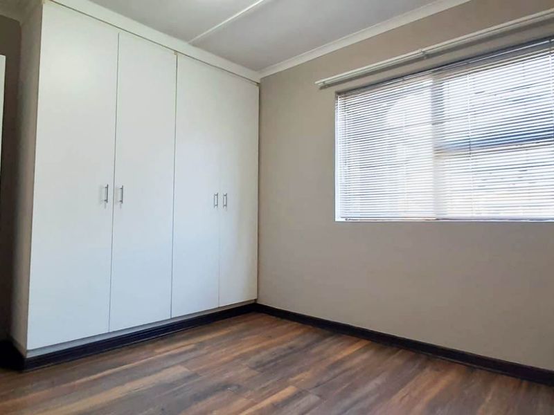A clean, empty bedroom featuring modern laminate flooring, built-in white cupboards, and venetian blinds, offering a neutral space ready for personalization.
