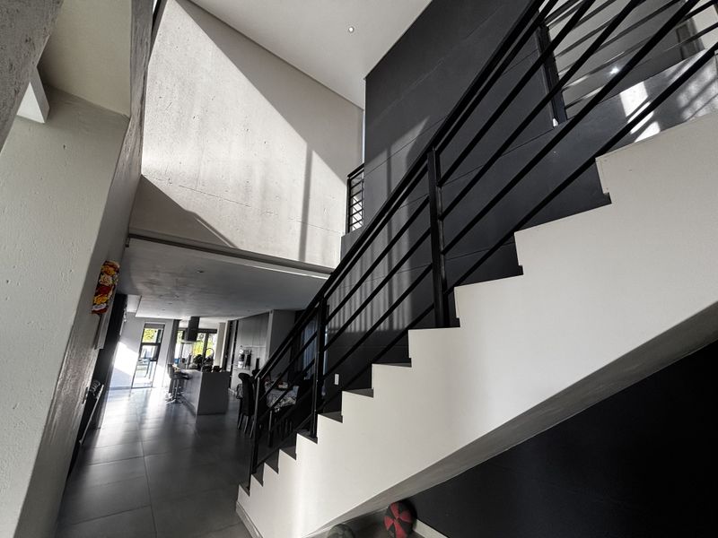 Modern open-plan living area featuring a striking staircase with metal balustrades, double volume ceilings, and sleek tiled flooring throughout.
