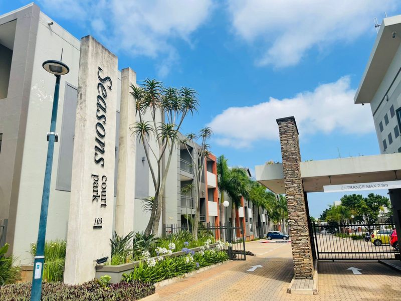 The entrance to Seasons Court apartment complex, featuring a secure gated access point, stone-clad pillars, paved driveway, and well-maintained landscaped gardens.