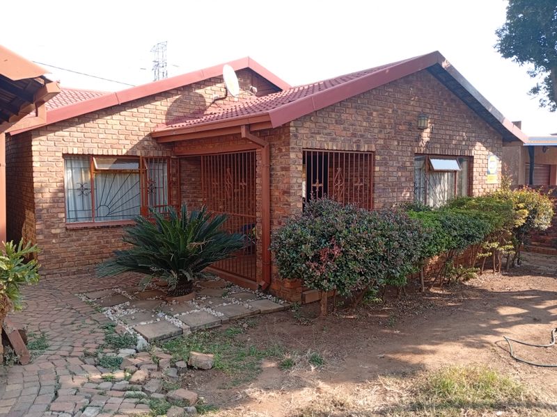 A well-maintained face brick house featuring a tiled roof, secure metal gates, and a paved front entrance area with established garden landscaping.