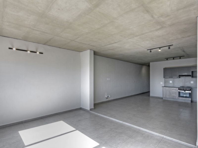 Spacious open-plan living area featuring an exposed concrete ceiling, modern tiled flooring, and a compact, functional kitchen with built-in appliances.