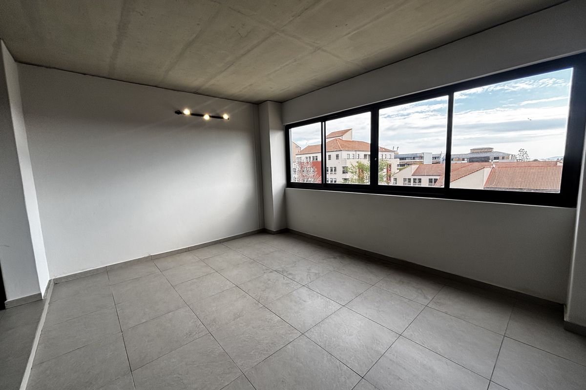 A bright, empty living area featuring modern tiled flooring, an exposed concrete ceiling, and large windows offering an urban view.