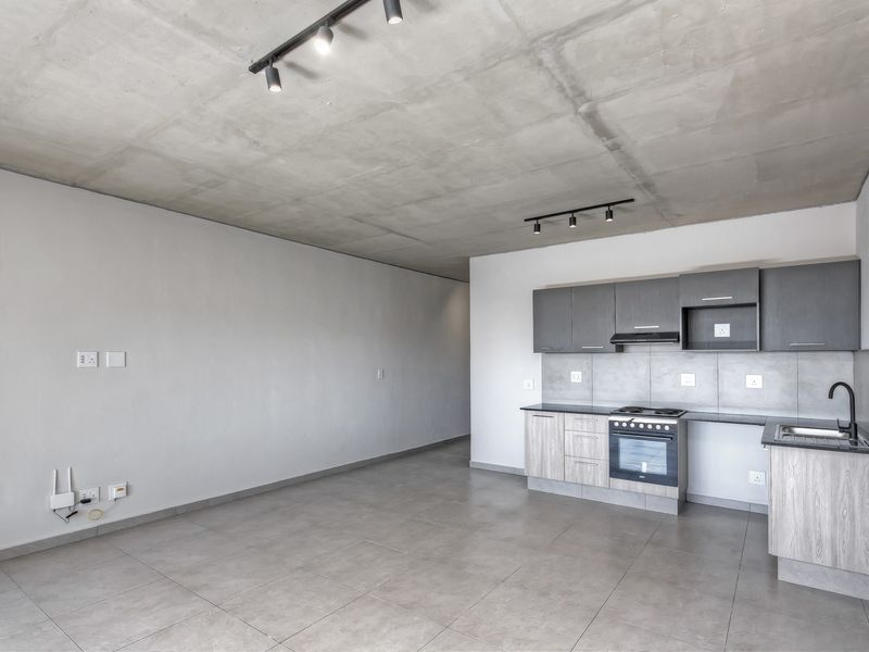 Modern open-plan living area featuring an exposed concrete ceiling, track lighting, tiled floors, and a compact kitchen with built-in cabinetry and appliances.