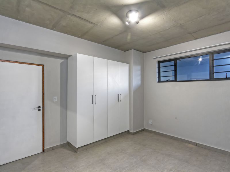 A clean, modern bedroom featuring an exposed concrete ceiling, tiled flooring, and a built-in white wardrobe, offering a neutral canvas for personalization.