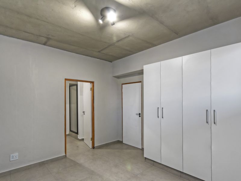 A clean, modern bedroom featuring built-in white cupboards, tiled flooring, and an exposed concrete ceiling, offering a neutral canvas for personalization.