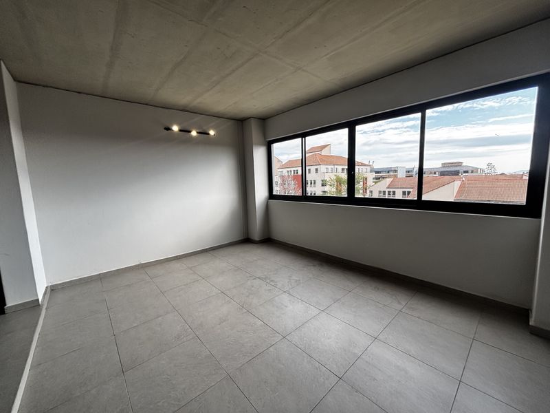A bright, empty living area featuring modern tiled flooring, an exposed concrete ceiling, and large windows offering an urban view.