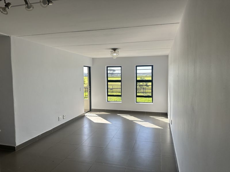 A bright, empty living area featuring neutral tiled flooring, clean white walls, and large windows providing natural light and views of the surrounding landscape.