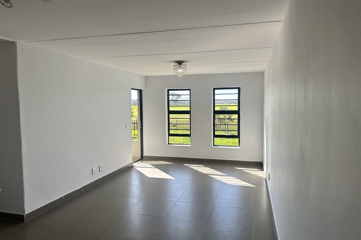 A bright, empty living area featuring neutral tiled flooring, clean white walls, and large windows providing natural light and views of the surrounding landscape.