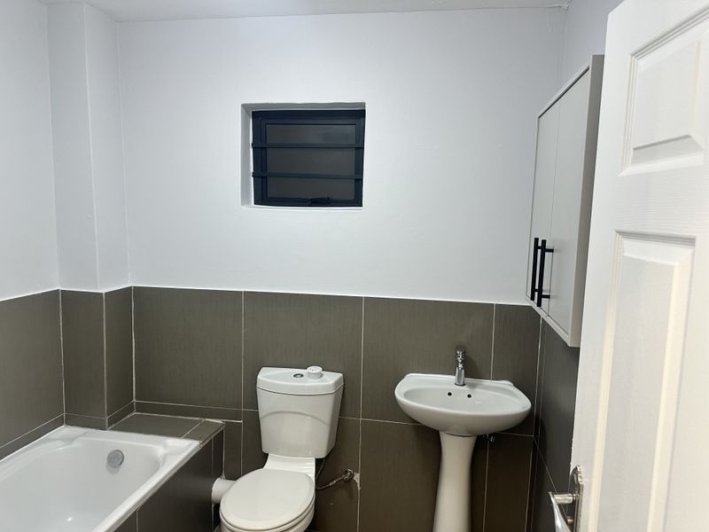 A clean, functional bathroom featuring a pedestal basin, toilet, and bathtub with neutral wall tiling and a small louvered window.