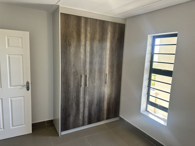 A clean, empty bedroom featuring modern wood-finish built-in wardrobes, tiled flooring, and a window fitted with security bars.