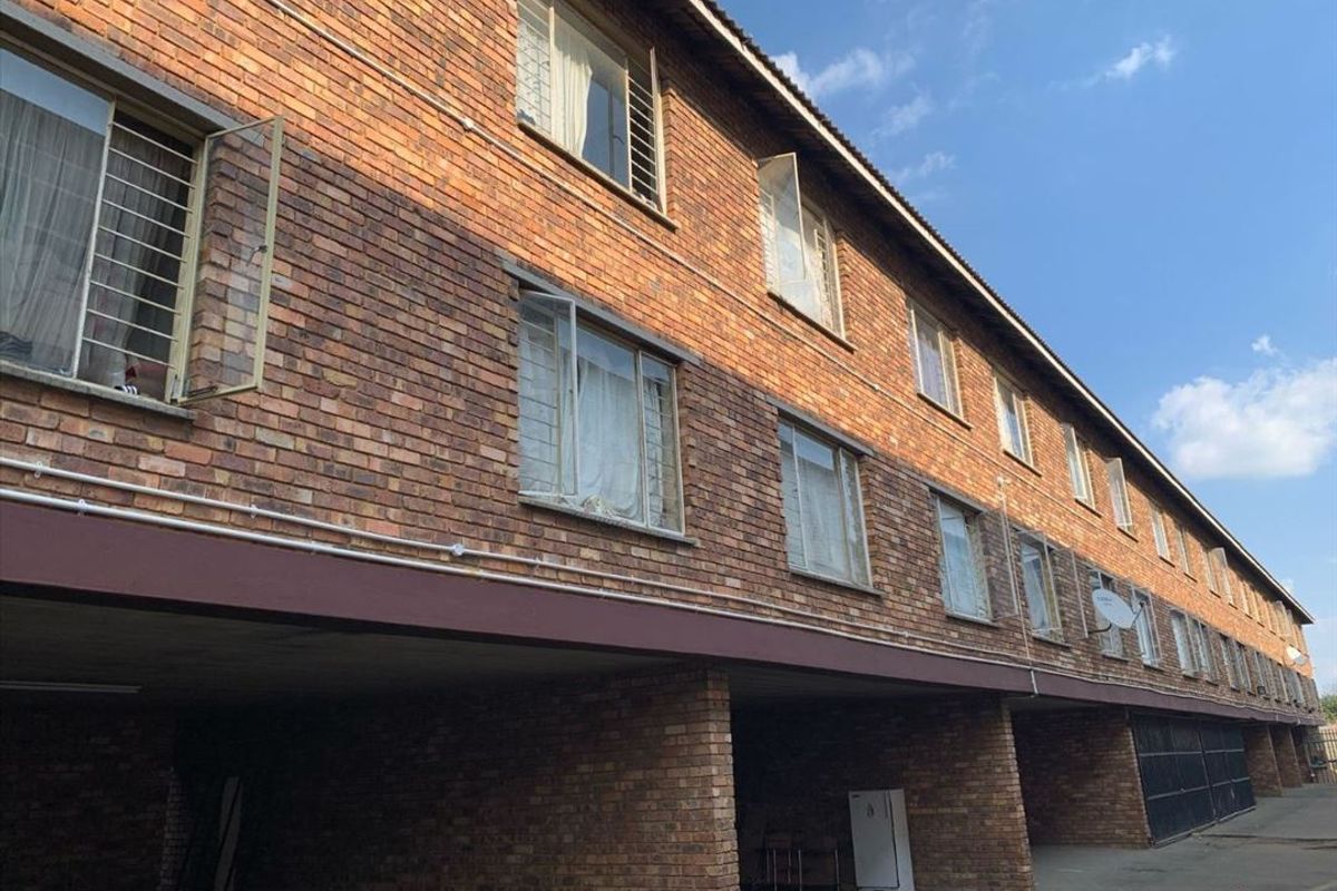 Exterior view of a multi-story apartment building featuring a face brick facade, casement windows with security bars, and a covered ground-level parking area.