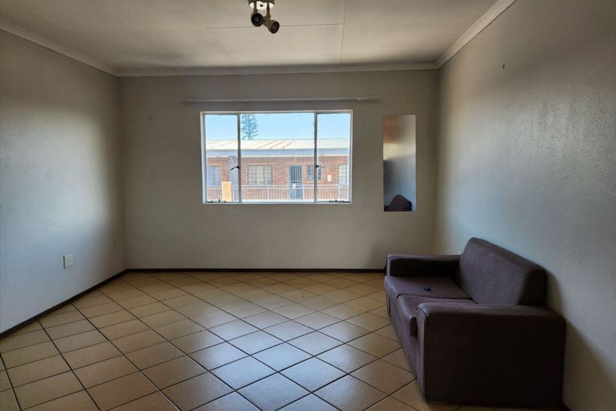 A spacious living room featuring ceramic tile flooring, neutral walls, and a large window providing natural light and views of the complex.