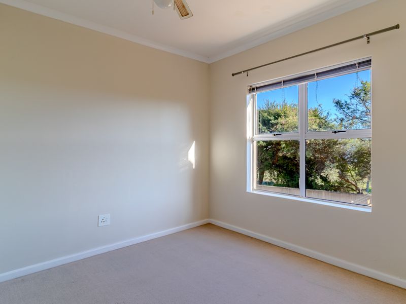 A bright, empty bedroom featuring neutral walls, carpeted flooring, a ceiling fan, and a large window with a curtain rod.