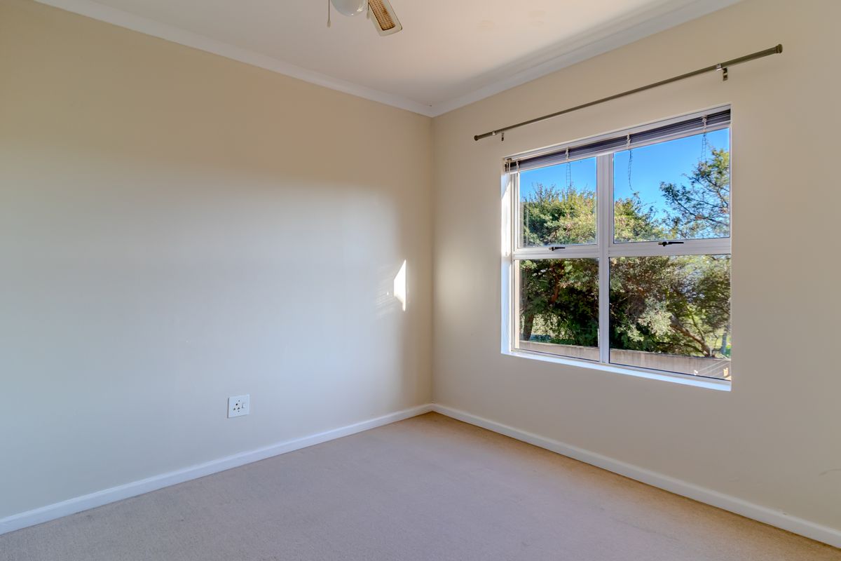 A bright, empty bedroom featuring neutral walls, carpeted flooring, a ceiling fan, and a large window with a curtain rod.