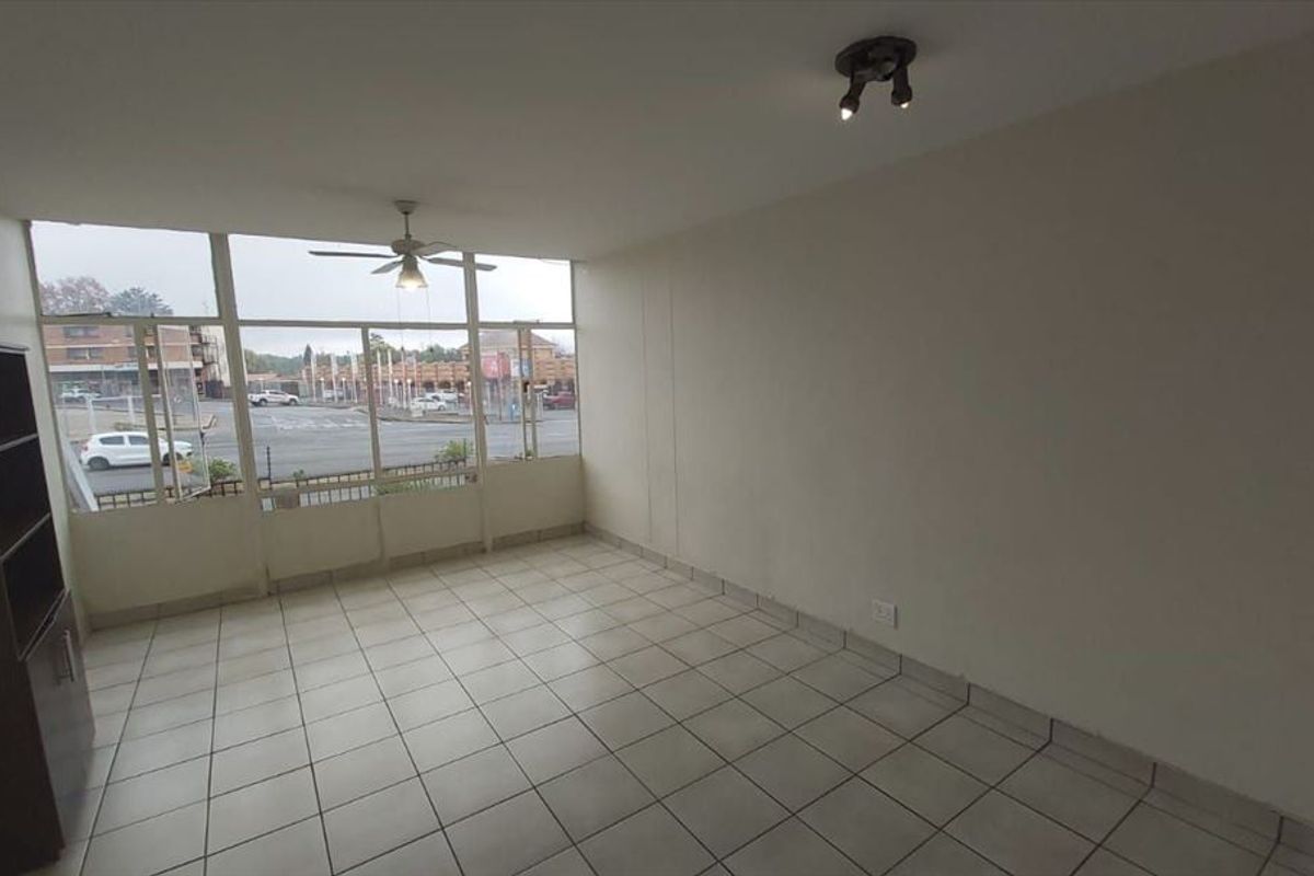 An empty, sunlit living area featuring tiled flooring, a ceiling fan, and large windows providing an exterior view.