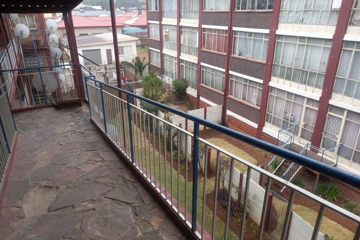An elevated balcony with stone flooring and metal railings, overlooking the apartment complex courtyard and surrounding residential area.