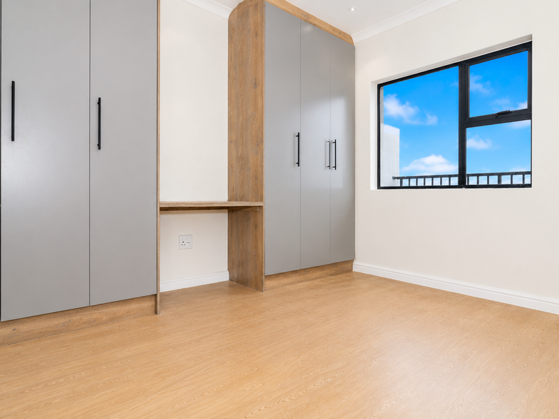 A bright, modern bedroom featuring light wood-look laminate flooring, ample built-in grey cabinetry with an integrated desk space, and a large window.