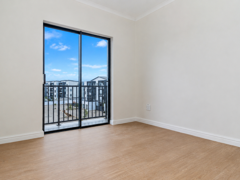 A bright, empty bedroom featuring modern laminate flooring, neutral walls, and a sliding glass door leading to a private balcony.