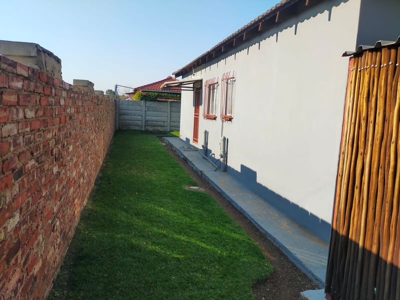 A side view of the property showing a narrow lawn area, a brick boundary wall, and the exterior wall of the house with security bars.