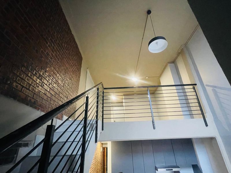 Open plan living area featuring a mezzanine level, exposed brick wall, and modern metal staircase railing, creating a spacious industrial-style interior.