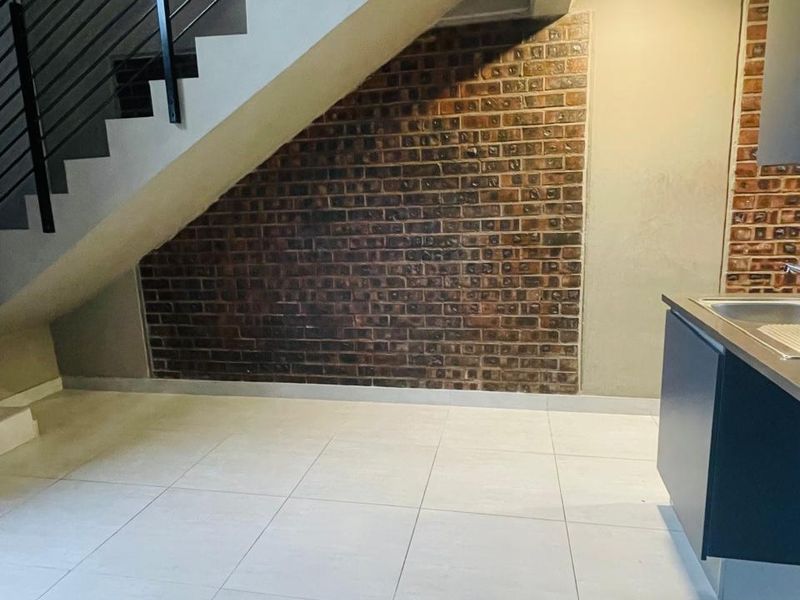 An open-plan living area featuring a modern staircase with a metal balustrade, an exposed brick feature wall, and tiled flooring with kitchen cabinetry.