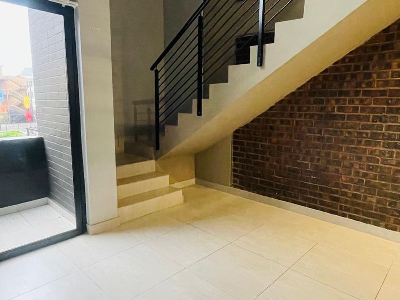 An open-plan living area featuring a modern staircase with metal balustrades, exposed brick feature wall, large format floor tiles, and access to a balcony.