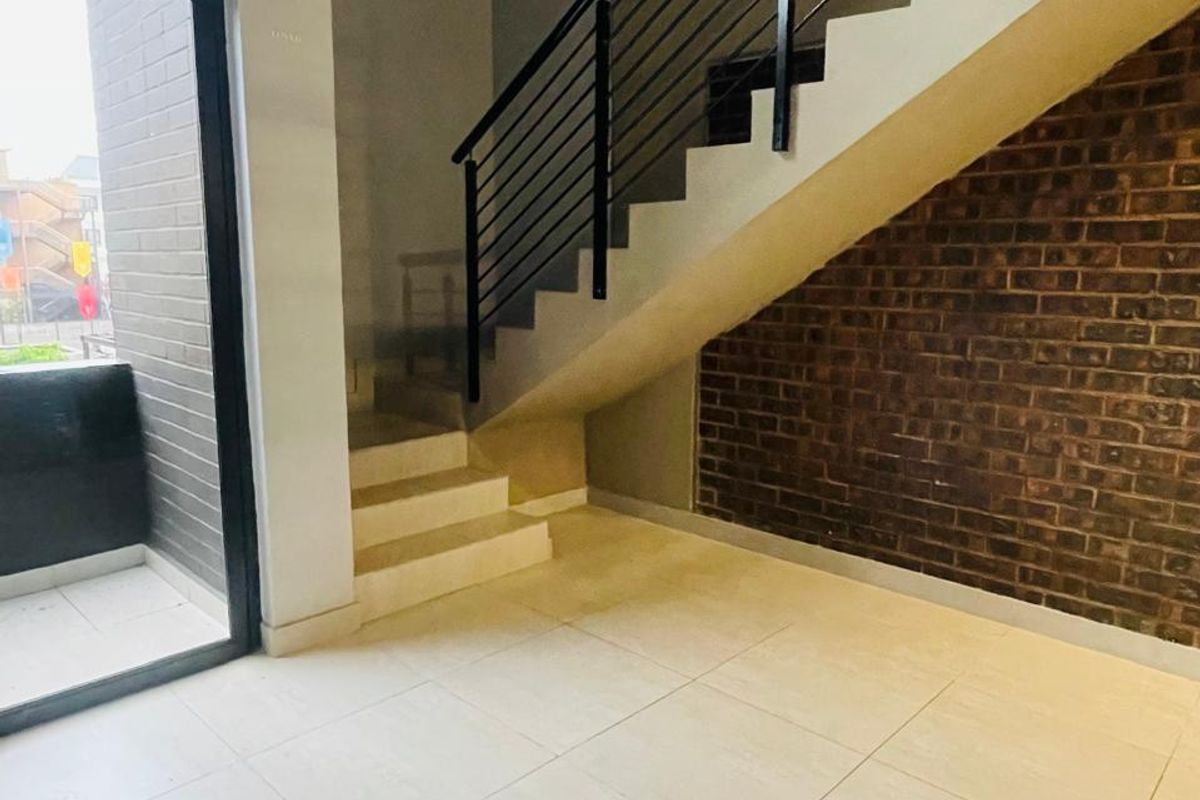 An open-plan living area featuring a modern staircase with metal balustrades, exposed brick feature wall, large format floor tiles, and access to a balcony.