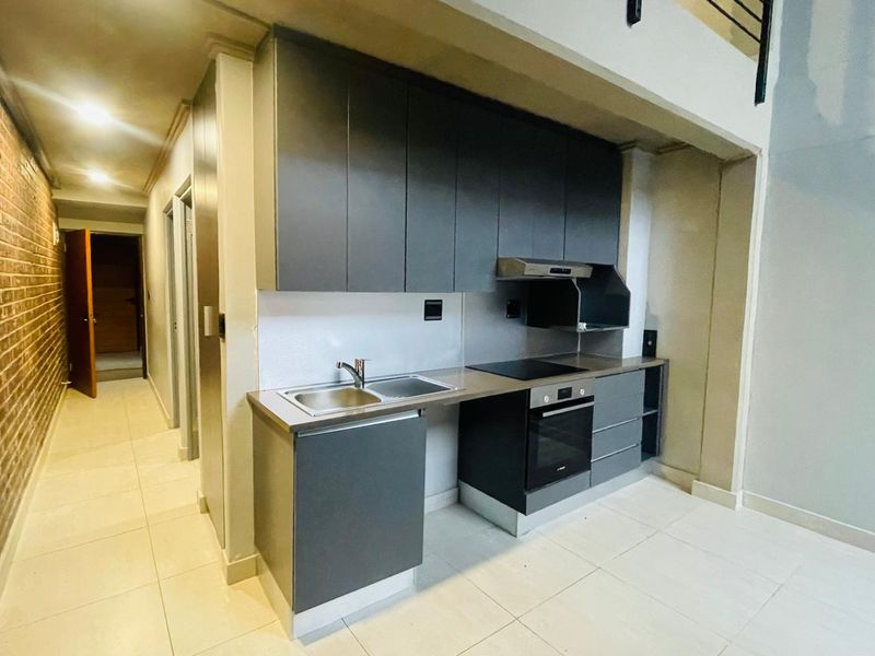 Modern kitchen featuring dark grey cabinetry, integrated oven and hob, stainless steel sink, and tiled flooring, set against an exposed brick feature wall.