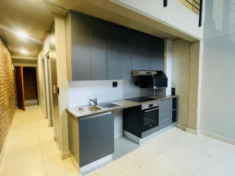 Modern kitchen featuring dark cabinetry, integrated oven and hob, stainless steel sink, and tiled flooring, set within an open-plan apartment layout.
