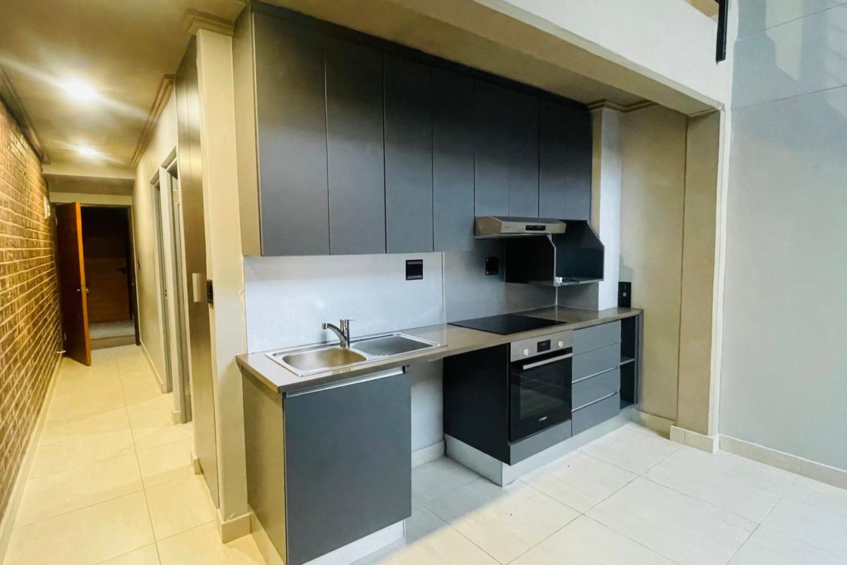 Modern kitchen featuring dark cabinetry, integrated oven and hob, stainless steel sink, and tiled flooring, set within an open-plan apartment layout.