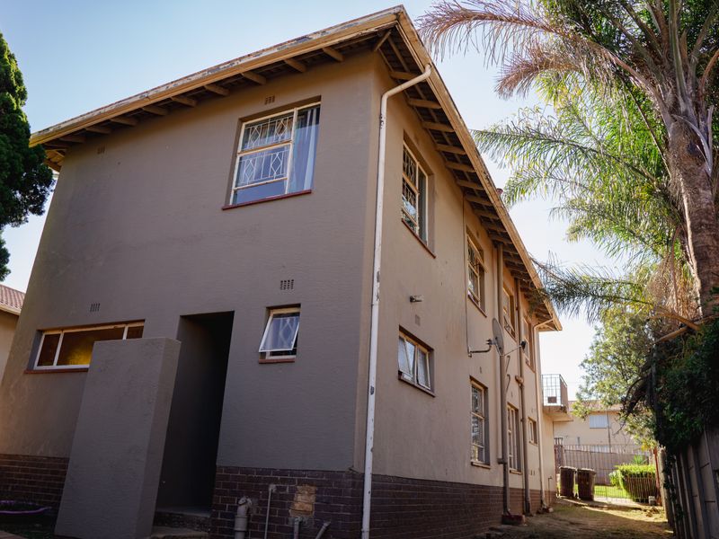 Side and rear view of a two-story residential building featuring plastered walls, a brick base, and multiple casement windows with security bars.