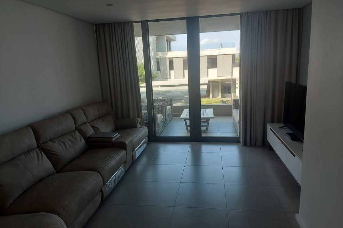 A modern living room featuring large sliding glass doors that open onto a private balcony, with neutral tiled flooring and recessed lighting.