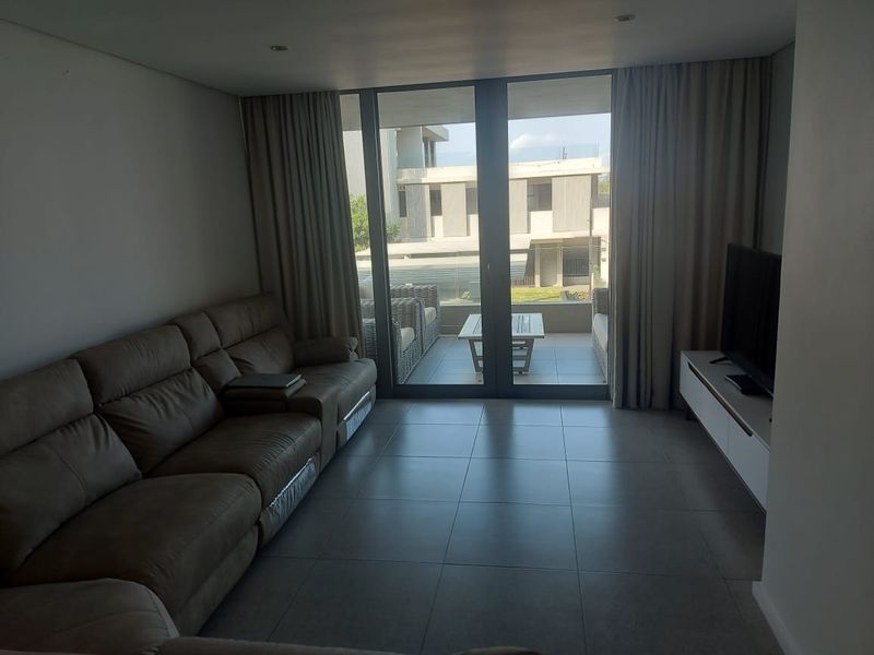A modern living room featuring large sliding glass doors that open onto a private balcony, with neutral tiled flooring and recessed lighting.