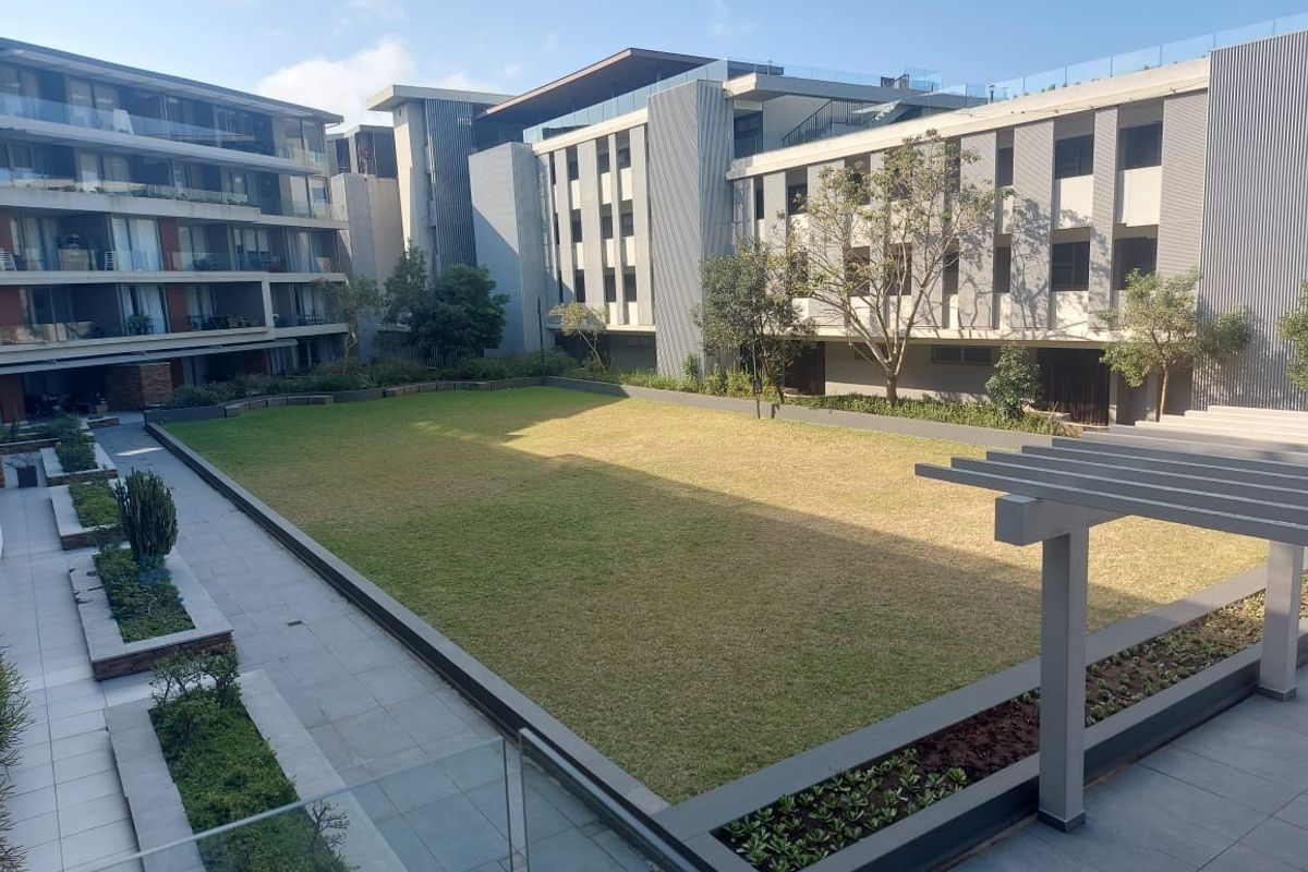 A well-maintained communal garden area featuring a central lawn, paved walkways, raised garden beds, and a modern pergola within an apartment complex.
