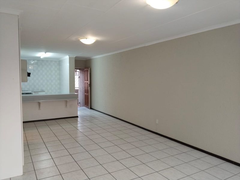 An open-plan living and kitchen area featuring neutral tiled flooring, a breakfast bar, and functional kitchen cabinetry in a clean, vacant space.
