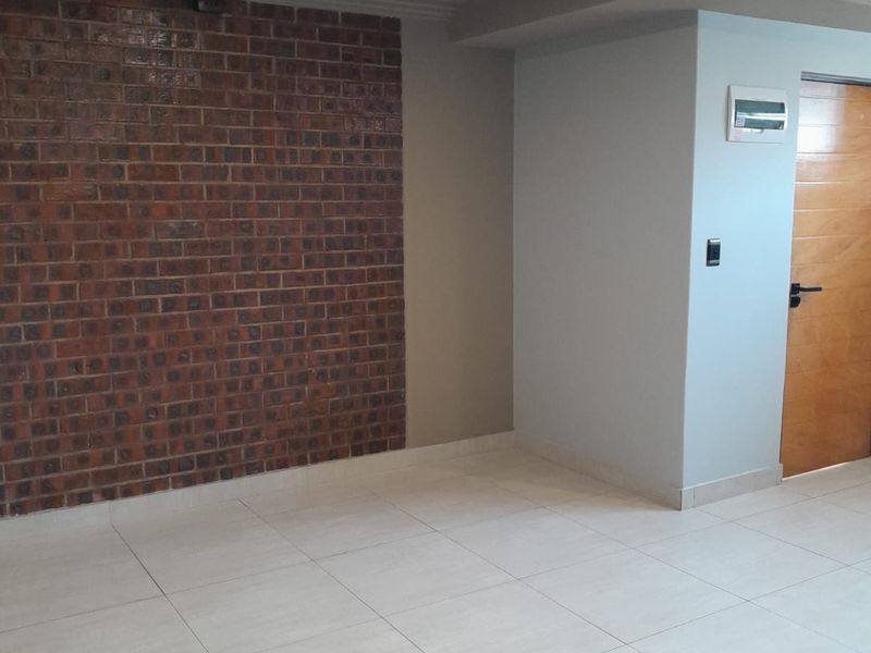An empty living area featuring a prominent exposed brick feature wall, neutral tiled flooring, and modern recessed lighting.