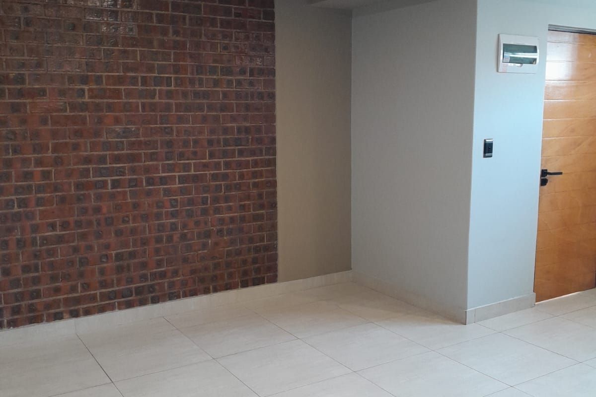 An empty living area featuring a prominent exposed brick feature wall, neutral tiled flooring, and modern recessed lighting.