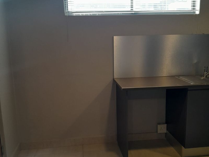 A compact kitchen area featuring a stainless steel countertop, integrated sink, and tiled flooring, illuminated by natural light through a window with venetian blinds.