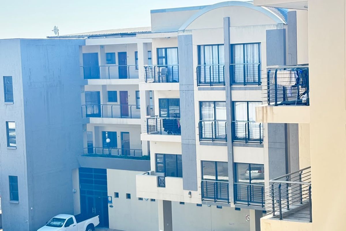 Exterior view of an apartment complex featuring multiple levels with balconies, metal railings, and a ground-level paved parking area.