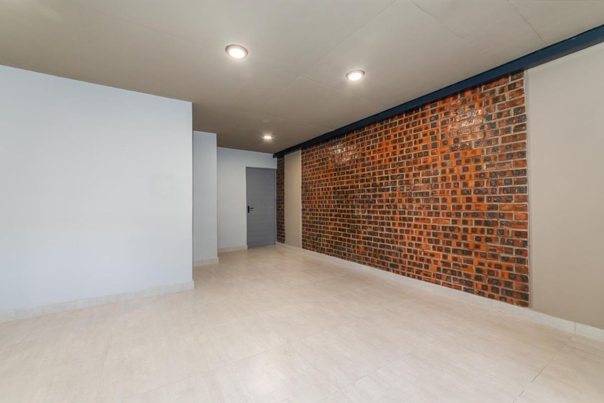 An empty, well-lit living area featuring a prominent exposed brick feature wall, neutral tiled flooring, and modern recessed ceiling lighting.