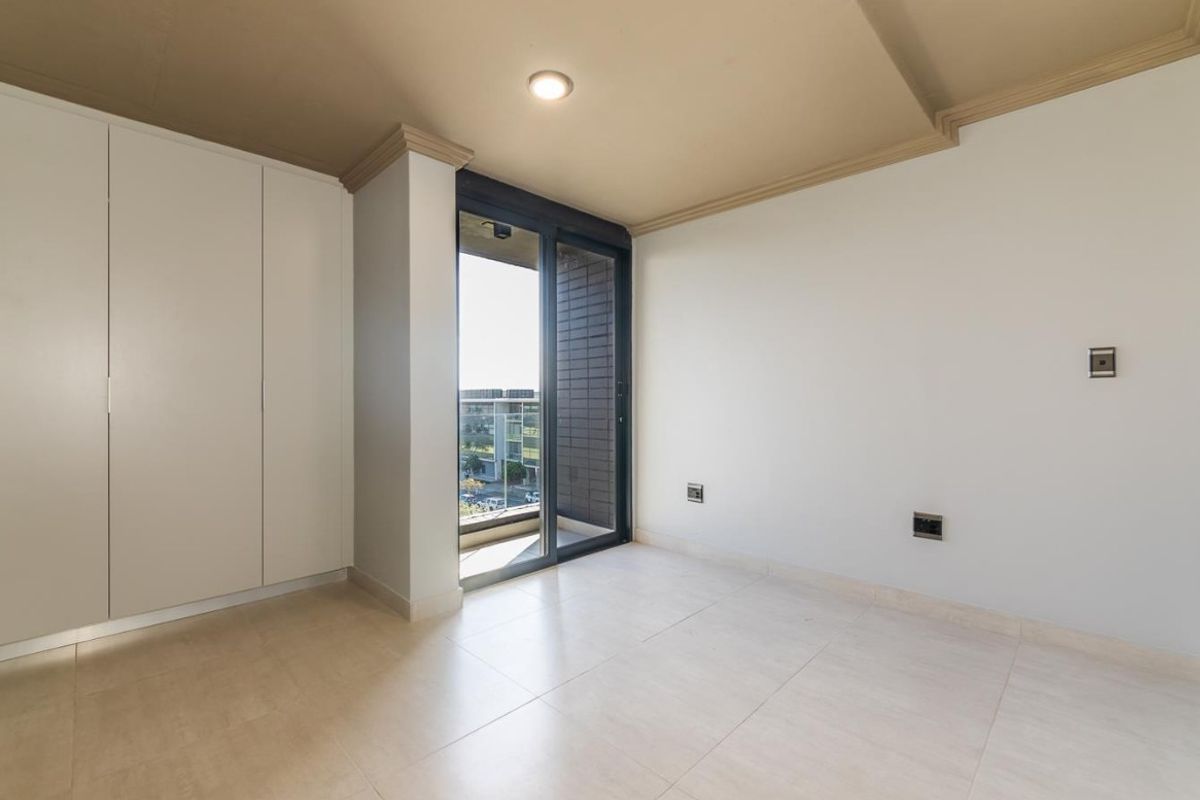 A bright, empty bedroom featuring built-in cupboards, tiled flooring, crown molding, and a sliding glass door leading to a balcony.