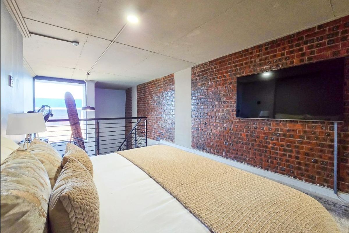 A modern industrial-style bedroom featuring an exposed brick feature wall, concrete ceiling, and a mezzanine-style layout with a metal staircase railing.