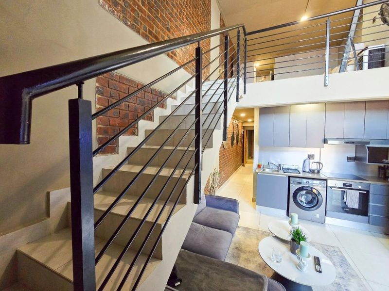 Modern open-plan apartment featuring a mezzanine level, exposed brick feature wall, contemporary kitchen cabinetry, and a sleek staircase with metal balustrade.
