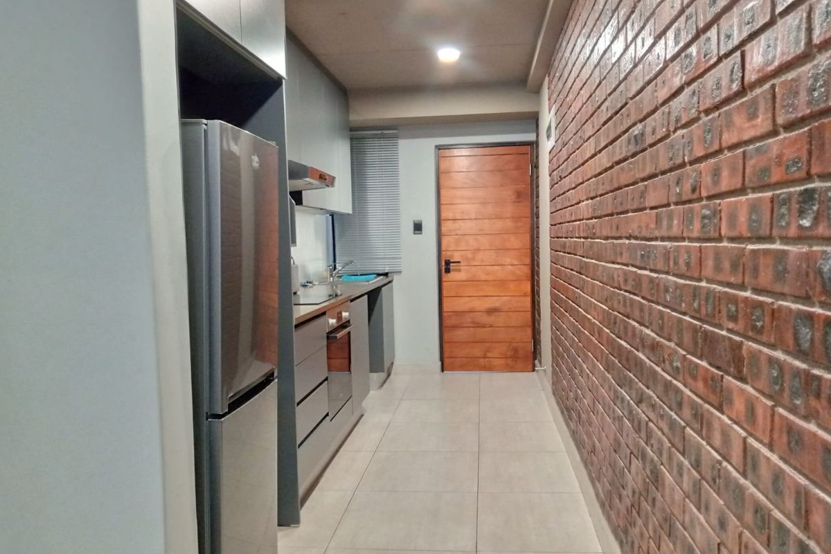 A modern galley-style kitchen featuring sleek cabinetry, integrated appliances, and a striking exposed brick feature wall, complemented by neutral floor tiles.