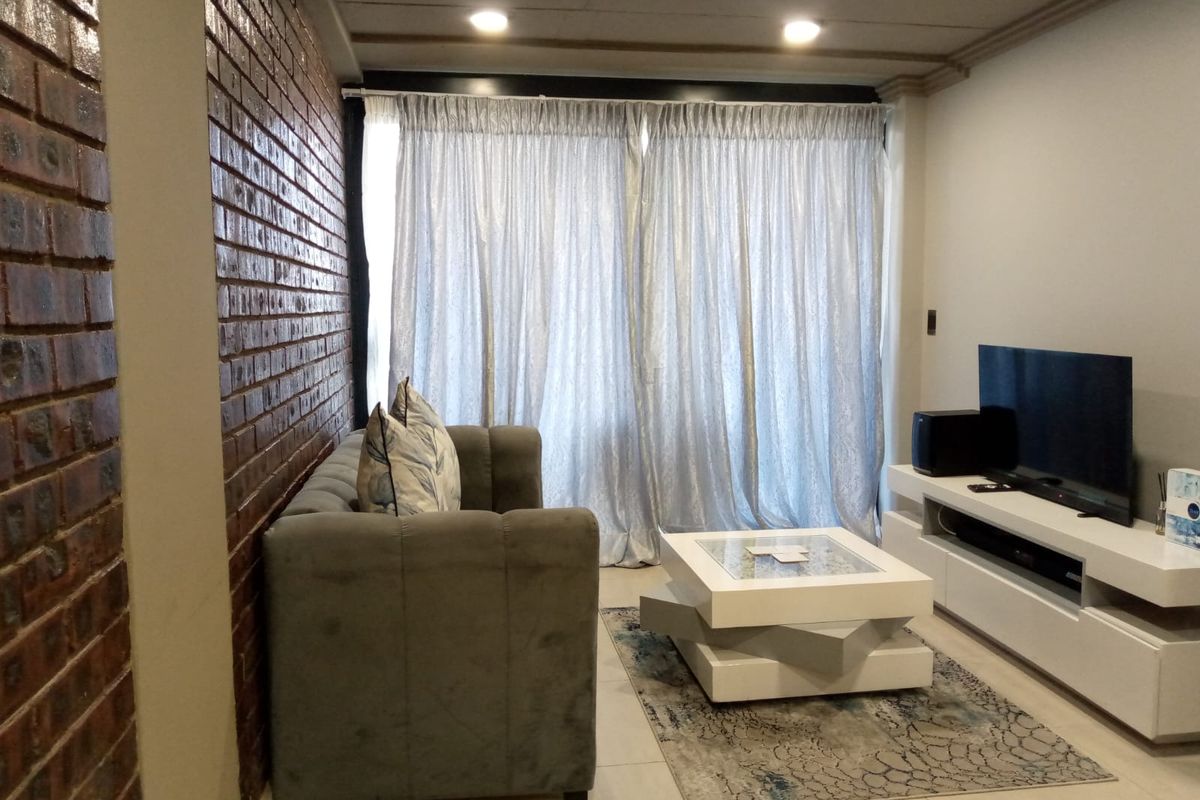 A comfortable living room featuring an exposed brick accent wall, tiled flooring, and recessed ceiling lighting, with access to a balcony through sliding doors.