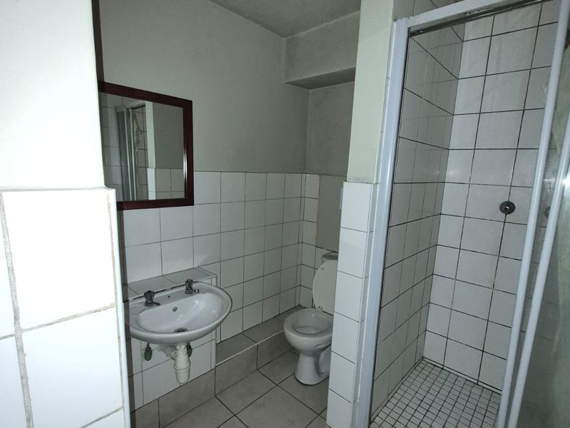 A functional bathroom featuring a shower enclosure, pedestal basin, and toilet with white ceramic wall and floor tiling throughout.